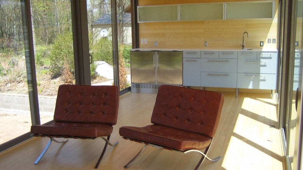对面的视野显示了厨房区域，白色和金属橱柜，玻璃门货架安装在墙上，前景是双皮革客厅椅。