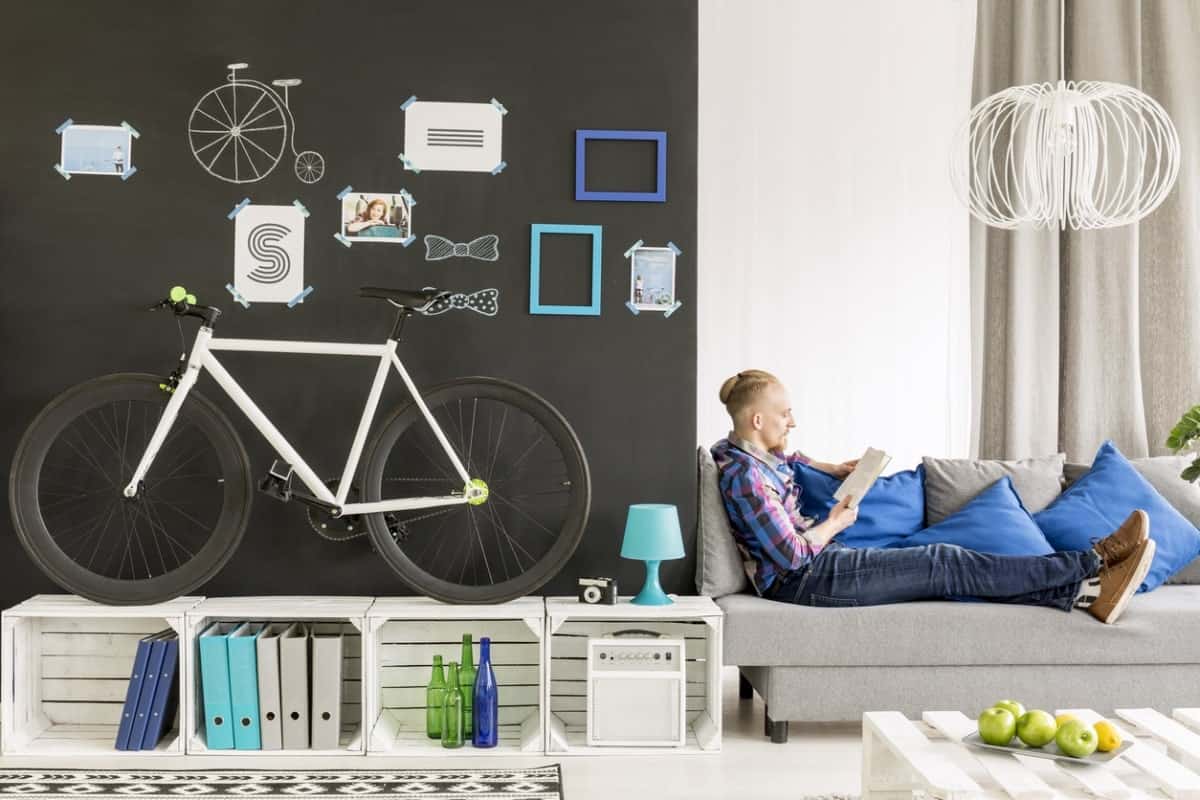 一辆自行车陈列在板条箱架子上，靠在客厅的黑色墙壁上，一个男人坐在沙发上看书。