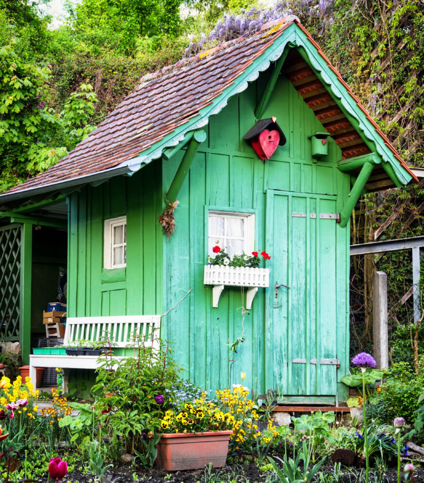 这座被漆成亮绿色的摇摇欲坠的花园小屋与周围的种植床相映成趣。一个小尖桩窗框和配套的长凳在明亮的绿色中格外显眼。