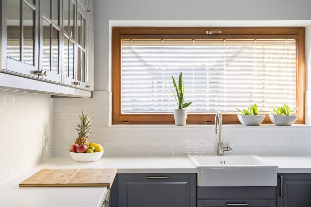 白色厨房，带木框窗户，盆栽植物，和一个农舍厨房水槽。