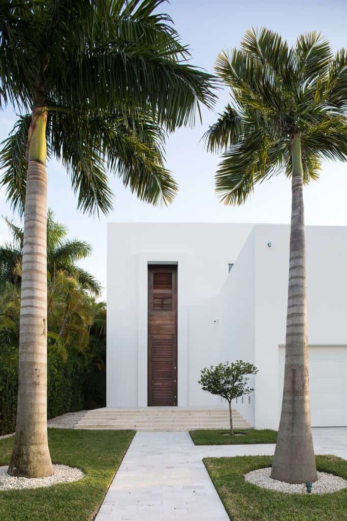 白色鹅卵石环绕的高大棕榈树突出了白色墙壁下的百叶窗前门。它软化了房子光滑、极简的设计。