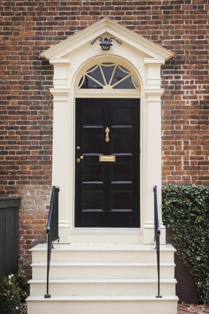 一扇简单的黑色门，由黄铜把手和门板制成。它由一个乳白色的门廊和一个配套的楼梯组成，与锻铁栏杆形成对比。