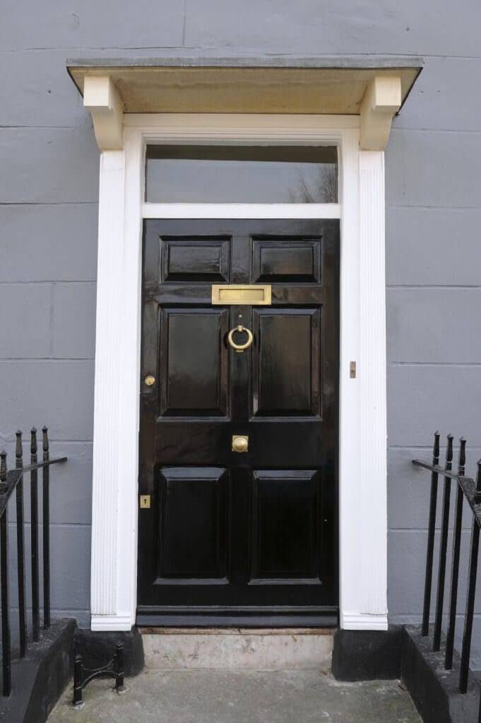 近距离观察这个黑色前门展示黄铜五金和锻铁栏杆。它周围环绕着白色的装饰，与灰色的混凝土墙相得益彰。