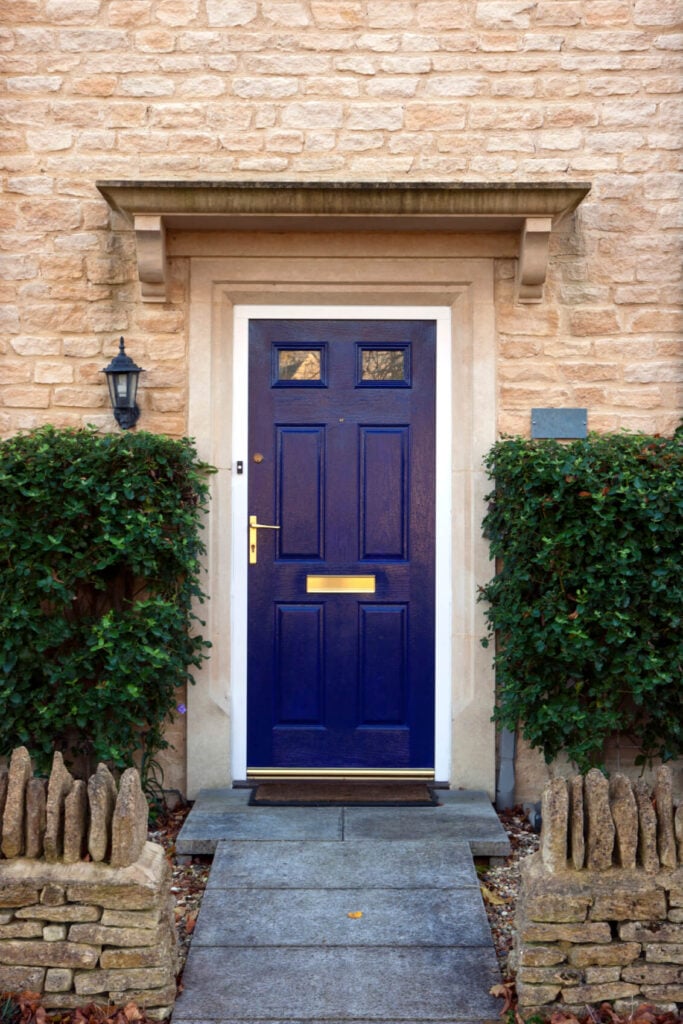 与上面的前门几乎相似，这扇蓝色的门配有黄铜把手和板，配以棕色镶边地毯和安装在石砖墙上的传统户外壁灯。