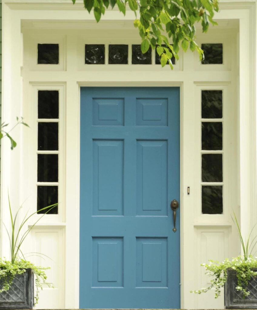 蓝色前门周围环绕着小玻璃板，门上装有青铜把手和把手。它在白色的墙壁和淡淡的绿色植物的映衬下格外显眼。
