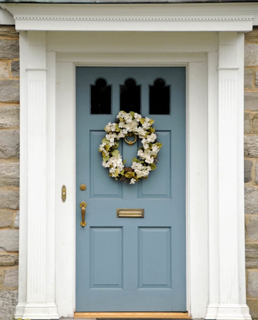白色的门廊与石砖墙相映成色，蓝色的门上装饰着一个可爱的花环。它完成了一个黄铜手柄和板以及一个环门环。