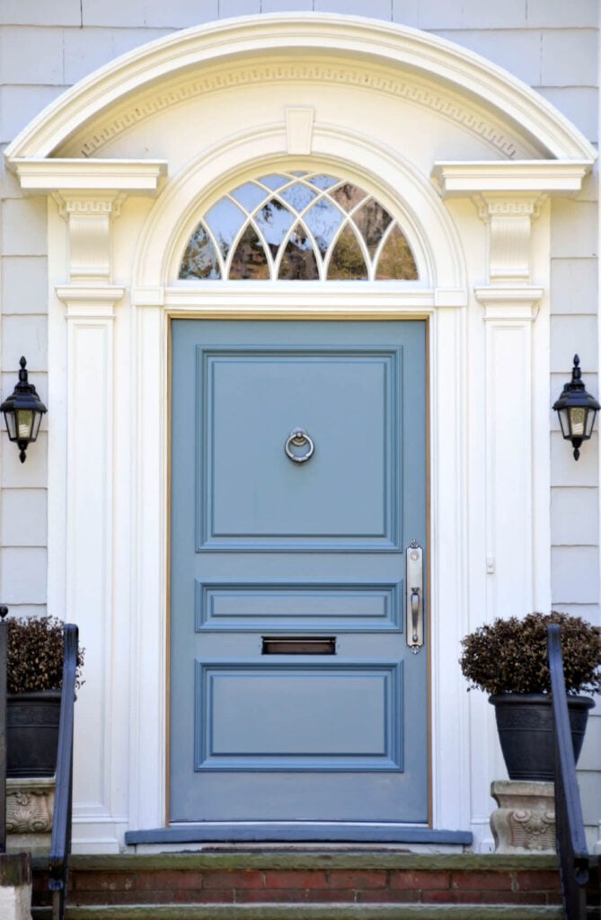 传统的户外烛台照亮蓝色的前门，内衬装有铬手柄和环门环。它的内衬是拱形装饰和白色的柱子。