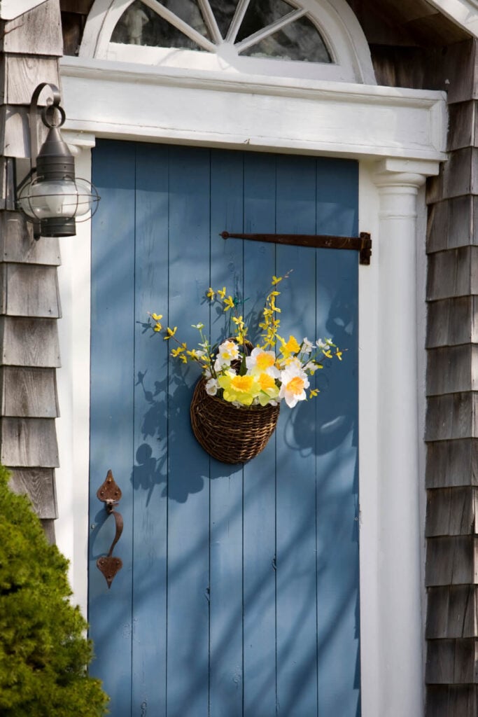 简单的蓝色前门由柳条植物支架装饰，与中性的壁板和白色柱子形成鲜明对比。它与金属烛台和旁边的新鲜植物搭配在一起。