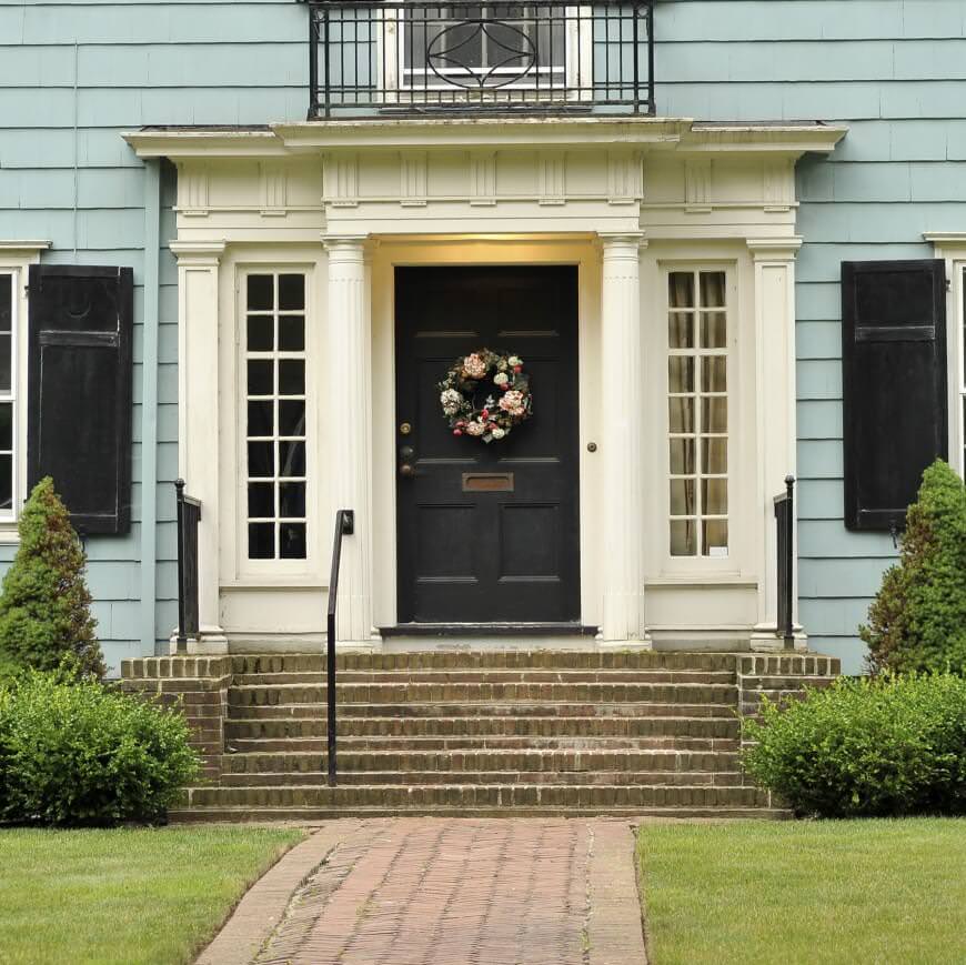 这座房子的特色是黑色的前门，上面装饰着一个华丽的花环。它配有白色的柱子和混凝土楼梯，楼梯两侧是郁郁葱葱的绿色草坪。