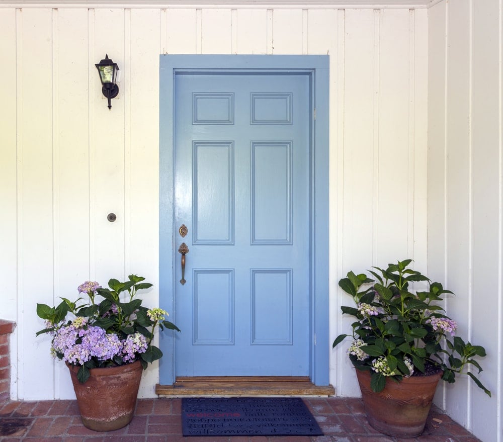 天蓝色的前门，黄铜把手，由安装在白色床头板上的壁灯照明。它配有盆栽植物和深蓝色地毯，铺在陶土地板上。