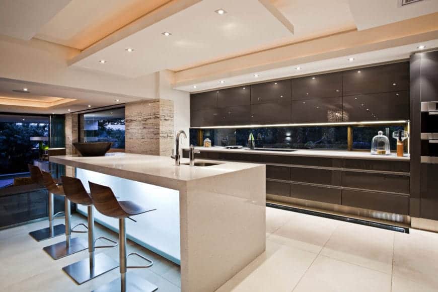 这是一个明亮而光滑的现代风格厨房，白色瀑布厨房岛与白色地板融为一体，并有发光的磨砂玻璃面板。它搭配了一个巧克力棕色的厨房半岛，内衬不锈钢细节。
