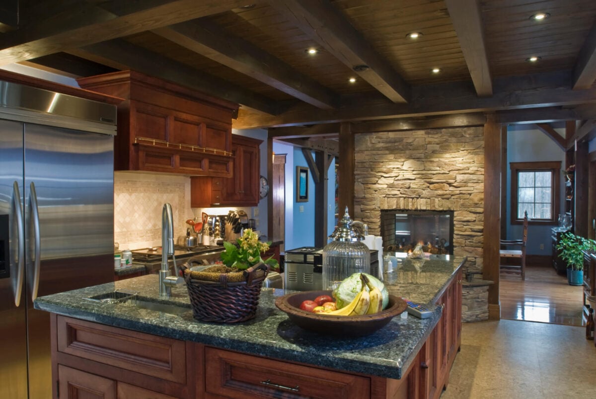 这是一间深色的厨房，厨房岛台上有深棕色的木制橱柜，烤箱和冰箱都放在木质结构中。深色的木质天花板与裸露的木梁相匹配，并与纹理石材壁炉相辅相成。