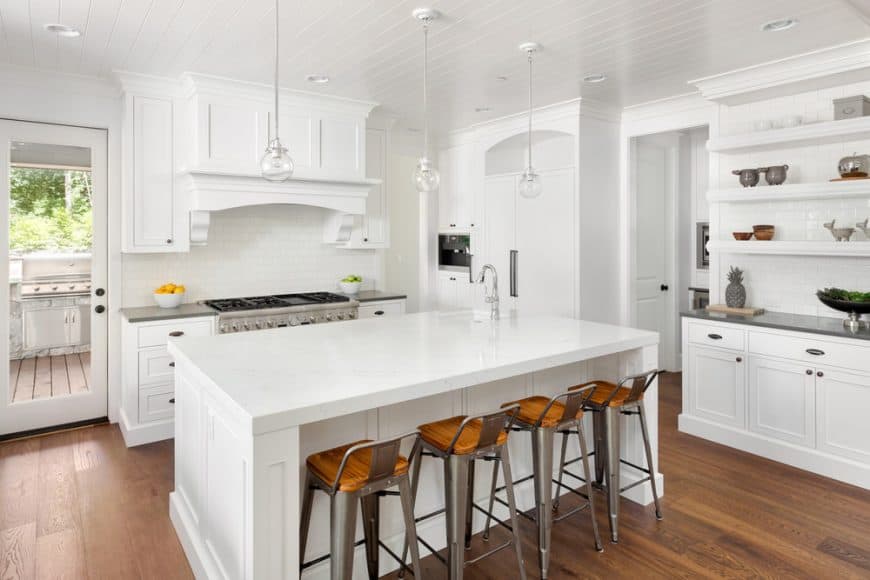 这个明亮的白色厨房的硬木地板与厨房岛的白色摇床橱柜和抽屉形成了很好的补充，周围的橱柜与白色的吊顶融合得很好。这种地板也与早餐吧的现代凳子搭配得很好。