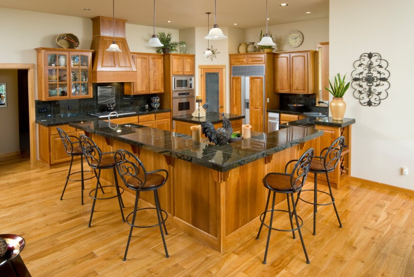 这个迷人的厨房的浅色硬木地板与两个木制厨房岛岛相匹配，融合在一起，厨房岛岛有一个黑色的台面，与棕色色调相辅相成，并作为配套的深色锻铁凳子的早餐吧台。