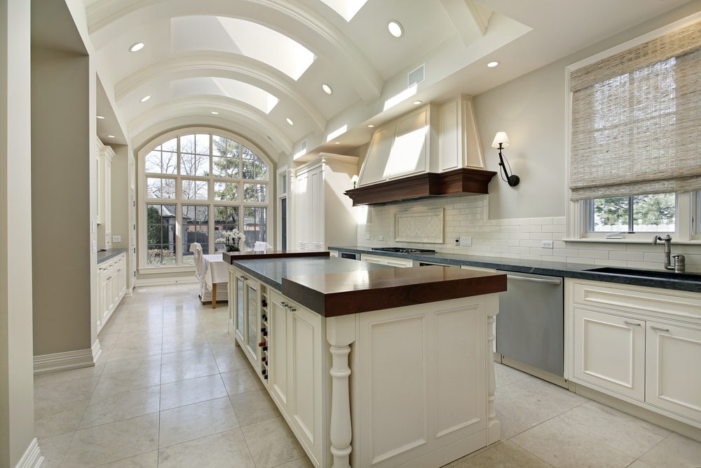 这是一个明亮的厨房，明亮的白色高天花板是拱形的，充满了天窗和嵌入式灯。这种亮度被带到大型厨房岛的白色摇床柜和抽屉上，以及墙上的橱柜上。
