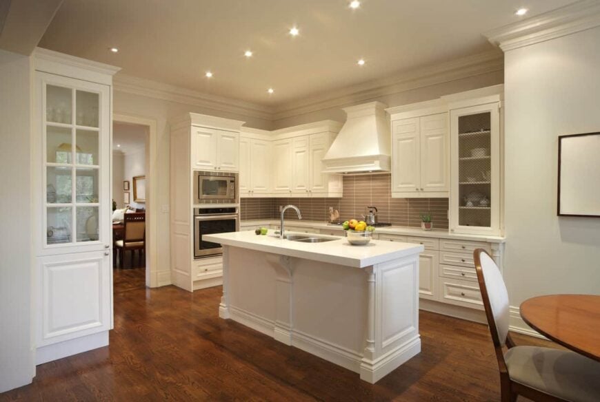 这间优雅厨房的明亮狭窄的厨房岛台在深色硬木地板的映衬下格外显眼。这种地板也与明亮的白色摇瓶柜和木结构的抽屉形成对比，木结构主导着墙壁，与灰色的后挡板形成平衡。