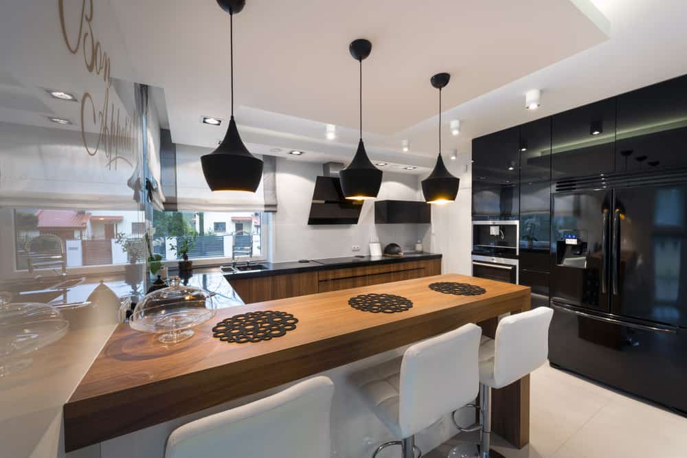 这个现代厨房的黑色电器赋予了它一种特殊的精致和奢华的美感。木制早餐吧台上方白色天花板上的黑色吊灯与白色的现代凳子形成了鲜明的对比。