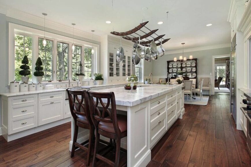 白色的厨房岛台顶部有一个弯曲的挂锅架，白色天花板上的凹槽灯使其更加明亮。这个架子与华丽厨房里的其他深色木质元素很搭配，比如木椅和硬木地板。