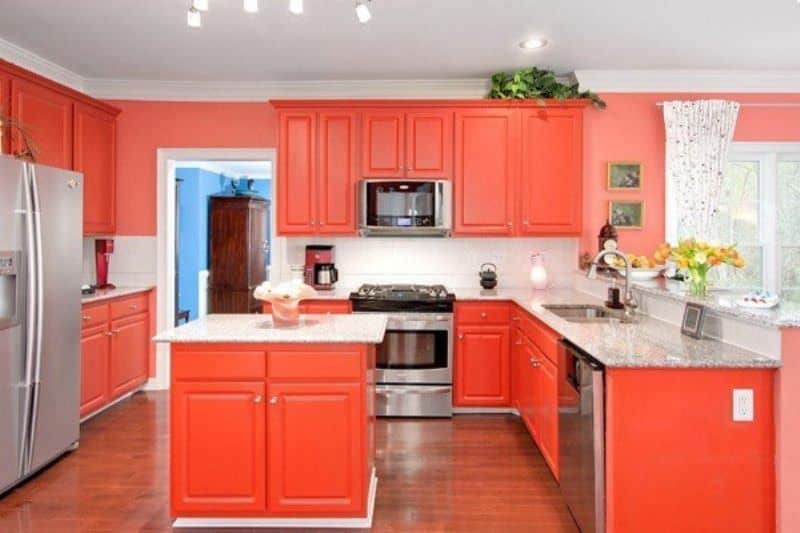 这是一个令人愉快的橙色厨房，有一个l形的半岛和一个小厨房岛，它的橱柜有同样明亮的橙色色调。这与墙壁的泥土橙色色调和硬木地板相匹配，硬木地板与白色天花板形成对比。