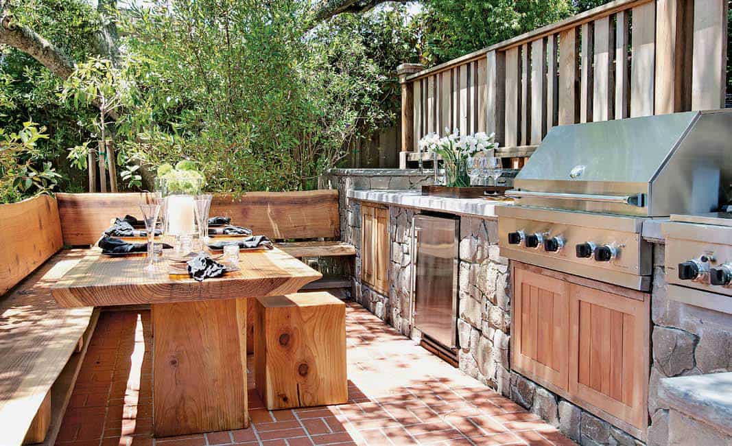 这是一个舒适的户外厨房，有一个裸露的木制餐桌的用餐角，内置的l型长凳和小凳子，与赤陶土地板非常相配。这也补充了不锈钢器具的纹理石材结构。