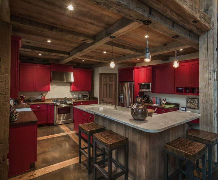 这个迷人而温馨的厨房的质朴木天花板与l型厨房橱柜的红色哑光橱柜形成了很好的木制补充，厨房橱柜有一个木制台面，以及角落里靠近灰色混凝土早餐吧的嵌入式办公桌。