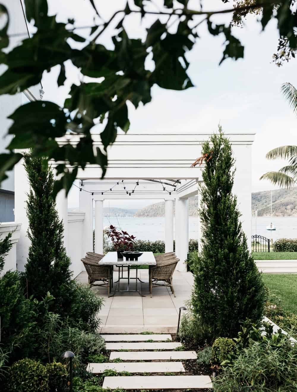 白色的庭院与美丽的小径被树木和绿色植物包围，通向户外餐桌。