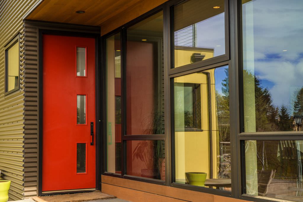 带玻璃插孔的光滑的红门和锻铁把手在这个现代化的房子里脱颖而出，展示了灰色的侧窗和玻璃镶板窗户。