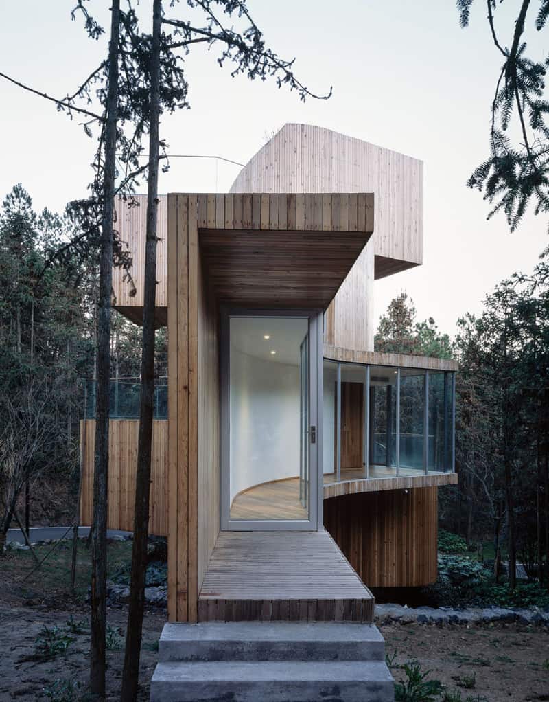 木材房屋展示平行的木板和弯曲的玻璃。它包括混凝土入口楼梯，通往木制平台，玻璃前门是铝制框架。