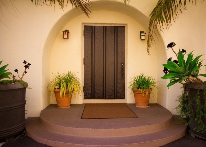 一个简单的木前门，在户外骗局照亮的拱形插图墙上。它是由绿色盆栽植物和一个混凝土着陆的棕色边毯互补。